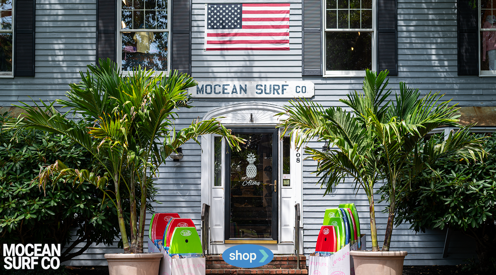 Front of a building with an American flag, 'Mocean Surf Co' sign, and colorful surfboards outside.